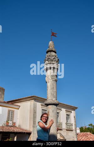 Una donna in pensione che indossa occhiali e una T-shirt verde chiaro appare sorridente e giocosa quando si "abbraccia" o si appoggia contro un'antica gogna, un'icona Foto Stock