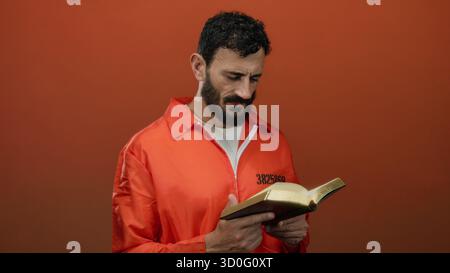 L'uomo ispanico con barba in tuta da prigione arancione tiene la bibbia su sfondo arancione, simboleggiando la riflessione e la redenzione. Foto Stock