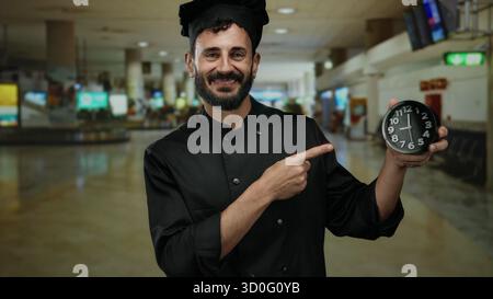 Lo chef ispanico in uniforme nera che punta all'orologio nel terminal dell'aeroporto evidenzia l'urgenza del tempo in mezzo a un ambiente di viaggio molto affollato. Foto Stock