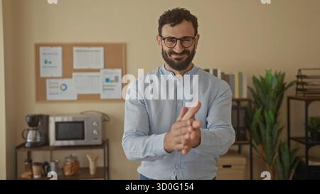L'uomo ispanico che indossa occhiali e barba applaude entusiasticamente in uno spazio ufficio con un microonde e documenti su una tavola da stiro. Foto Stock