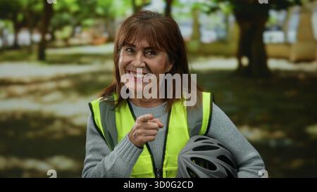 Una donna anziana ispanica sorride in un parco mentre punta la macchina fotografica, indossa un giubbotto riflettente e tiene in mano un casco da bicicletta, incarnando la sicurezza durante un Foto Stock