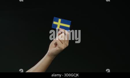 Mano che regge una bandiera svedese su sfondo nero, simboleggiando la nazionalità e l'orgoglio svedesi, isolata e prominente nella scena, capturin Foto Stock