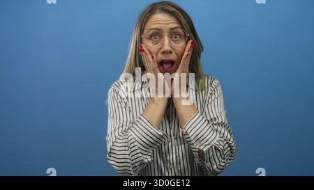 Donna che indossa occhiali rotondi tiene le mani alle guance in un ambiente da studio blu con una blusa a righe; sorpresa. Foto Stock