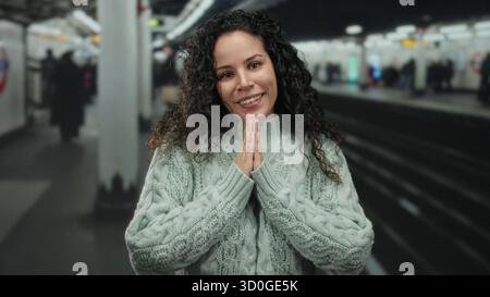 Donna sorridente in stazione ferroviaria con maglione con le mani in posa di preghiera, che suggerisce gratitudine in un ambiente ferroviario affollato. Foto Stock