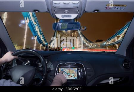 Guidare un'auto mentre si utilizza il touch screen di un sistema di navigazione GPS verso il Tower Bridge di notte, simbolo di Londra, Regno Unito Foto Stock