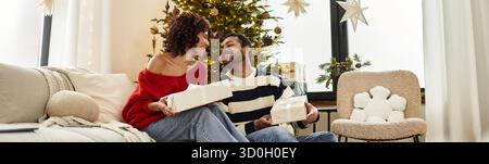 Una coppia felice condivide risate e scambi di regali con il loro albero di Natale splendidamente decorato, striscione Foto Stock
