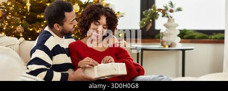 Una coppia gioiosa celebra il Natale, scambiandosi regali con il loro albero splendidamente decorato, lo striscione Foto Stock
