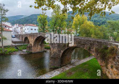 Un antico ponte ad arco in pietra attraversa un fiume tranquillo in un pittoresco villaggio, con case bianche e tetti in terracotta annidati contro lussureggianti colline boscose Foto Stock