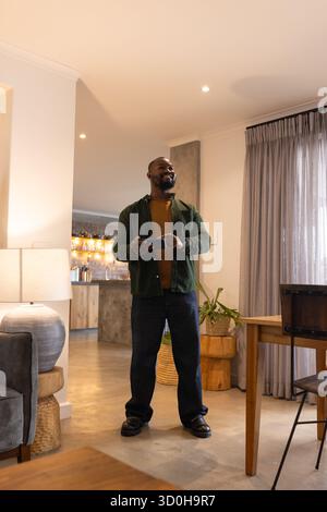 Uomo afroamericano in piedi con smartphone in una spaziosa area soggiorno con bancone bar e impianto Foto Stock
