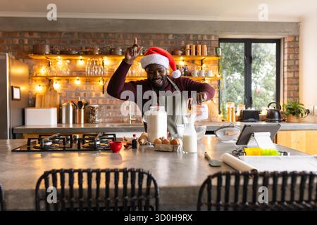 Uomo afroamericano che indossa il cappello di Babbo Natale e il grembiule verde che misura la farina sull'isola in cucina rustica Foto Stock