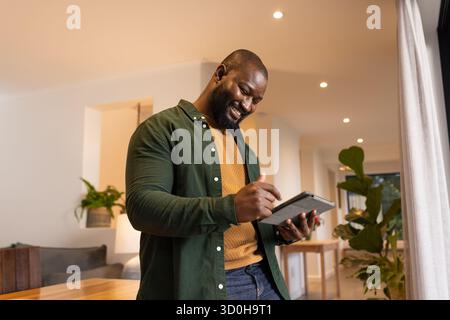 Uomo afroamericano in piedi in salotto a scrivere su tablet con stilo e maglione senape Foto Stock