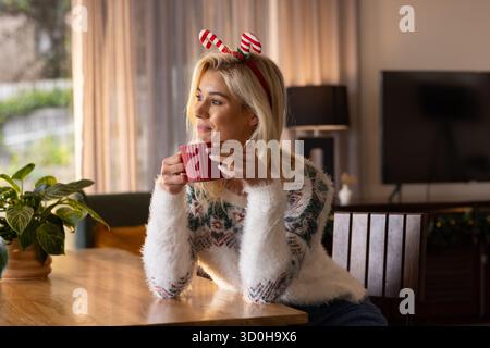 Donna appoggiata sul tavolo da pranzo a casa con un maglione natalizio, fascia a righe che regge una tazza rossa Foto Stock