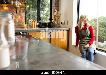 Donna che indossa grembiule verde, maglione rosso seduto sul davanzale in cucina e che guarda il giardino con tostapane Foto Stock