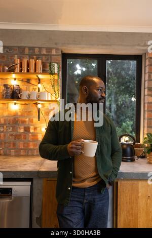 Uomo afroamericano appoggiato al bancone nella cucina di casa che tiene la tazza bianca vicino al bollitore nero Foto Stock