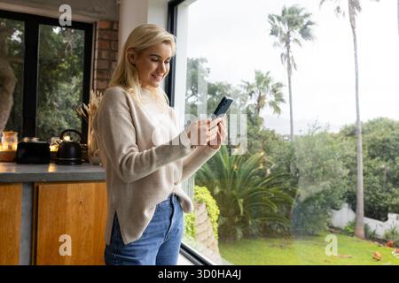 Donna di 20 anni in piedi davanti a una finestra alta della cucina che regge lo smartphone con vista sul giardino, copia spazio Foto Stock