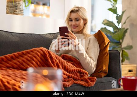 Donna adulta seduta sul divano grigio di casa con smartphone con coperta arancione e lume di candela Foto Stock