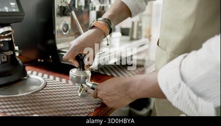Il barista indossa un grembiule di taupe che tampona i fondi nel portafiltro alla barra del caffè con il macinacaffè e la lancia del vapore Foto Stock