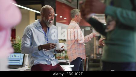 Uomo barbuto sorridente che indossa una camicia azzurra che tiene una tazza di caffè e un piattino in un ufficio open space Foto Stock