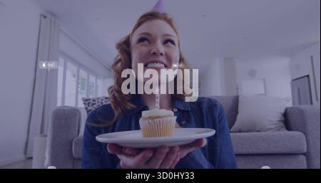 Donna sorridente che indossa una camicia in denim che regge un cupcake con una candela accesa a casa, con icone dei social media Foto Stock