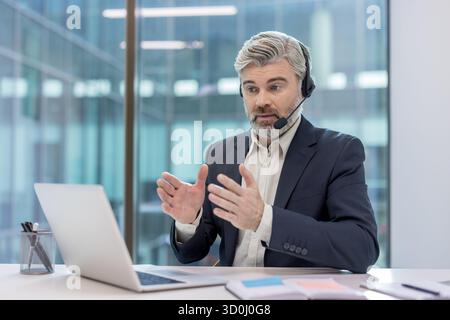 Uomo d'affari che ha una riunione virtuale, comunica con clienti o colleghi mentre lavora in remoto da un ufficio moderno, fornendo supporto e discutendo attivamente Foto Stock