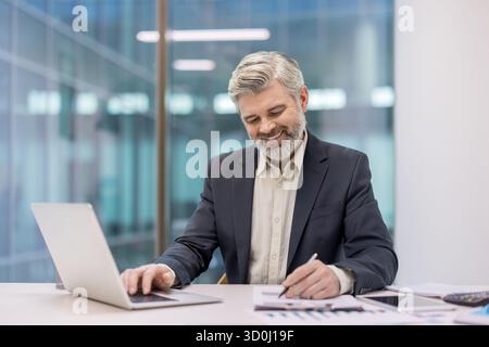 Uomo d'affari anziano che lavora alla scrivania dell'ufficio, digita su un computer portatile e scrive appunti sui documenti, sorridendo durante la revisione di grafici e dati finanziari Foto Stock