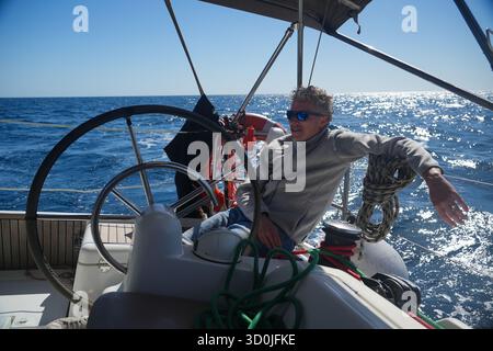Uomo rilassato con occhiali da sole al timone di uno yacht a vela in mare aperto in una giornata di sole, trasmettendo libertà, svago e stile di vita di viaggio. Foto Stock