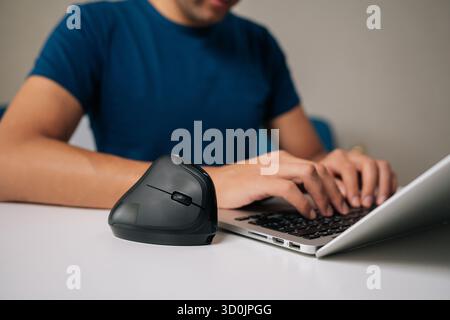 Foto ritagliata di un professionista di successo che lavora su una scrivania bianca minimalista con mouse verticale ergonomico, scrivendo sul laptop in una casa moderna e luminosa Foto Stock