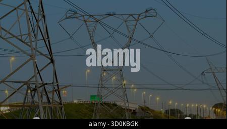 Torre in traliccio che sostiene le linee elettriche lungo l'autostrada al crepuscolo, con pali della lampada e cartello verde Foto Stock