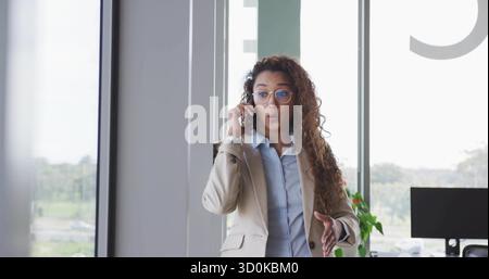 Donna d'affari che parla con smartphone e gesti accanto alla finestra dell'ufficio con un impianto di monitoraggio da scrivania Foto Stock