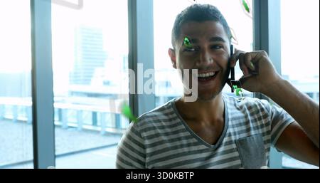 Uomo sorridente in una maglietta casual che tiene in mano lo smartphone che parla vicino alle finestre dell'ufficio con grafica e spazio di copia Foto Stock