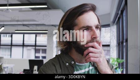 Uomo barbuto che indossa una maglietta verde e una camicia verde oliva pensando in ufficio, con monitor e vetrine Foto Stock