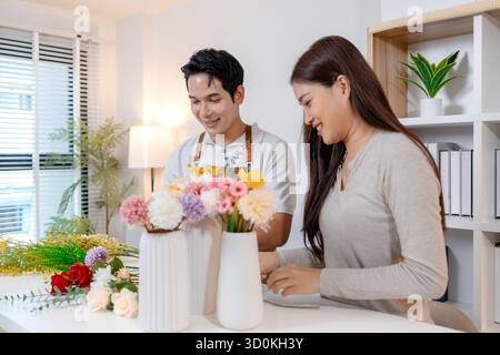 Una giovane coppia che organizza fiori colorati insieme in un ambiente accogliente. L'uomo, indossando un grembiule, aiuta la donna a creare un bellissimo bouquet, shar Foto Stock