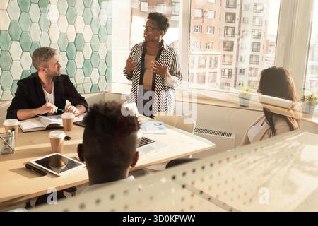 Elegante donna afroamericana che parla alla riunione d'affari in una moderna sala conferenze, colleghi che la ascoltano attentamente Foto Stock