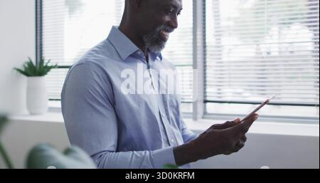 Uomo sorridente che indossa una camicia azzurra che tocca il tablet sul davanzale dell'ufficio, con succulente e persiane Foto Stock