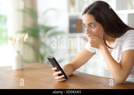 Donna scioccata che controlla contenuti sorprendenti sul cellulare di casa Foto Stock