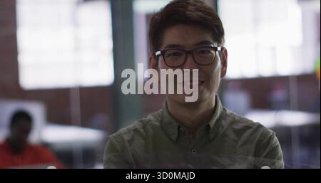 Uomo asiatico sorridente che indossa camicia verde e occhiali alla scrivania dietro una partizione di vetro con computer portatile Foto Stock