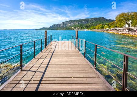 Veduta di un moderno molo in legno che si estende sulle acque blu del Lago di Garda nel villaggio di Garda, in Italia. Passeggiata sul lago con ristoranti nel backgrou Foto Stock
