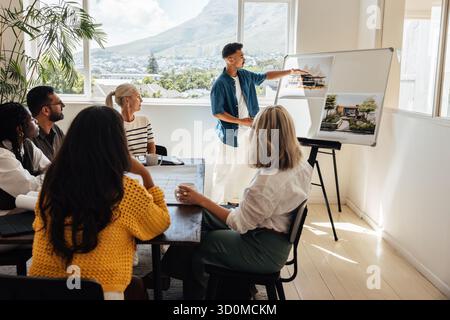 Giovane architetto che presenta i concetti di design ai colleghi in un ambiente di riunione collaborativo in studio. Il team include diverse persone impegnate in un Foto Stock