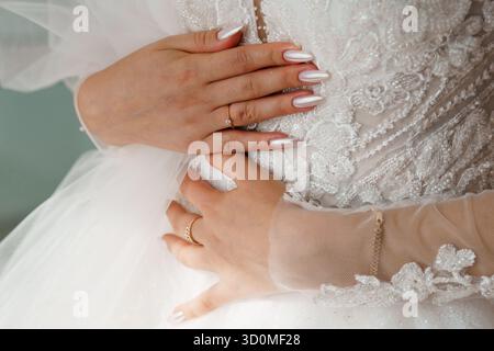 Un primo piano cattura le mani delle spose adornate da delicate fedi nuziali e bracciali dorati contro l'intricato pizzo del suo abito da sposa bianco, evoki Foto Stock