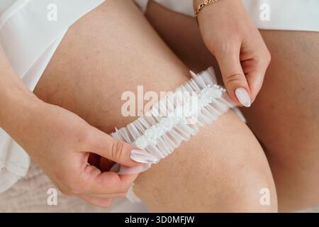Uno scatto ravvicinato raffigura una sposa che applica con cura una giarrettiera in pizzo bianco decorata con delicati dettagli di perle durante i preparativi per il matrimonio, catturando inti Foto Stock