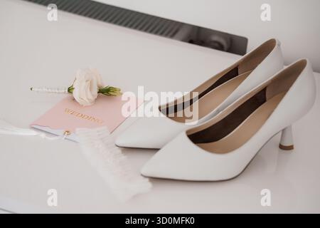 Uno scatto ravvicinato di décolleté da sposa in raso bianco con un'unica rosa blush e un invito a nozze rosa, a significare l'eleganza della sposa e la gioiosa anticipazione Foto Stock