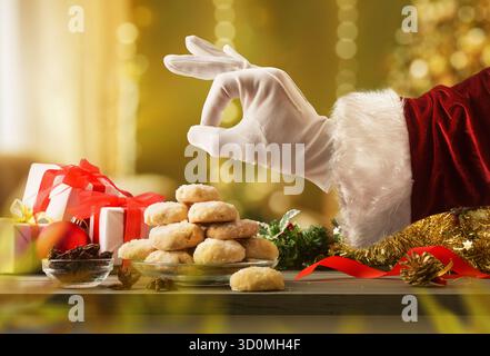 La mano di Babbo Natale con un buon gesto e tavolo con una pila di biscotti all'anice e zucchero decorati con elementi natalizi e un caldo sfondo dorato Foto Stock
