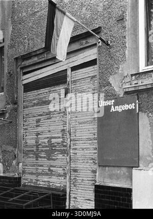 DEU, Germania, Berlino, 1990-03-18: Un negozio di alimentari in rovina nel quartiere Heinersdorf del quartiere berlinese di Pankow, appena un quarto di anno dopo l'apertura del muro di Berlino. Le tende del negozio sono abbassate, la tavola accanto all'ingresso con la scritta "Unser Angebot" (la nostra offerta) è vuota. Una bandiera tedesca è appesa sulla facciata in brollizione. HF [traduzione automatizzata] Foto Stock