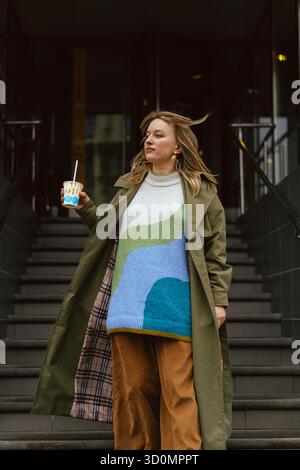 Donna elegante che regge un drink sui gradini urbani indossando un trench all'oliva Foto Stock