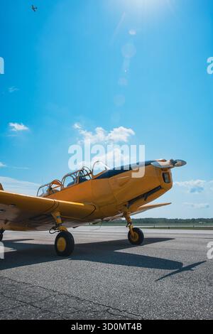 Velivolo da addestramento giallo d'epoca restaurato parcheggiato sull'asfalto sotto un cielo estivo blu. La luce del sole crea un bagliore dell'obiettivo nell'inquadratura, mentre un secondo Foto Stock