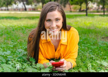 Giovane donna sorridente sdraiata sull'erba che tiene in mano lo smartphone e guarda la fotocamera Foto Stock
