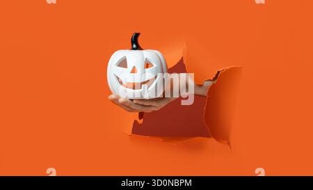 Jack-o'-Lanterna bianca in mano. Una Jack-o'-Lantern in ceramica bianca con stelo nero è trattenuta da uno sfondo arancione lacerato. Foto Stock