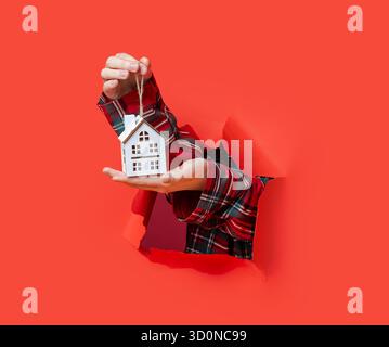 Buon Natale. Le mani reggono un piccolo ornamento bianco in legno per la casa di Natale con una corda attraverso la carta rossa strappata. Foto Stock