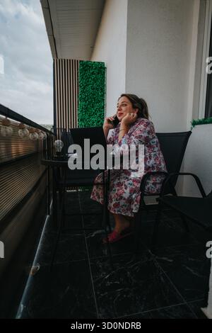 La donna con abito floreale lavora su un computer portatile mentre parla al telefono sul balcone dell'ufficio domestico Foto Stock
