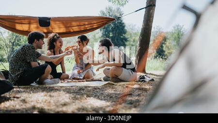 Gli amici condividono la pizza durante una soleggiata giornata di campeggio sotto un telo con un accogliente picnic Foto Stock